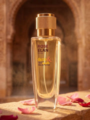 A 50ml bottle of ROSE ÉLAN Unisex Eau de Parfum by dazze & blussh, featuring a clear glass bottle filled with golden-amber liquid and a textured gold cap, set against a warm, blurred stone archway background with scattered pink rose petals.